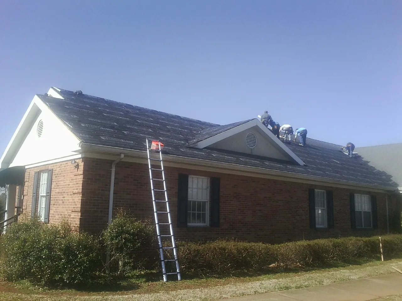 Crew tearing shingles off a brick ranch roof with a ladder set — Emergency Roof Repair work by our crew in Dunbarton