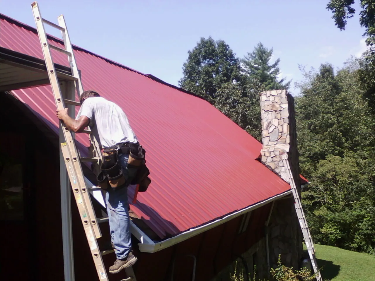 Roofer on a ladder beside a red metal roof and stone chimney — Soffit & Fascia Repair by Roofing Brothers in Dunbarton